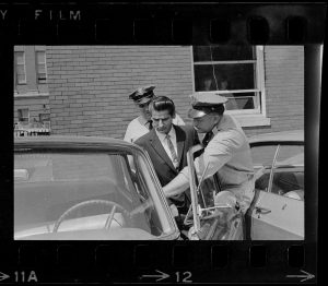 Positive image of a photographic negative showing two police officers placing a man in an automobile.